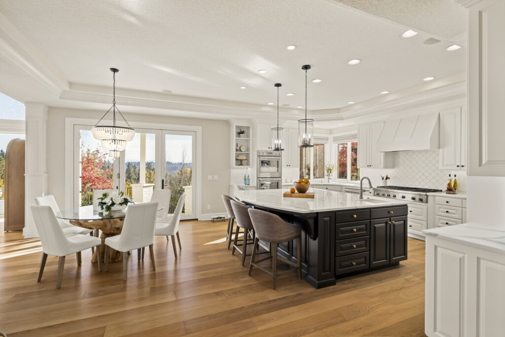 Kitchen and dining area showcasing a spacious layout with white cabinetry, a large black island with seating, and hardwood floors. Large windows and glass doors provide natural light and a view of autumn foliage outside, while pendant lights and a chandelier add elegant lighting.