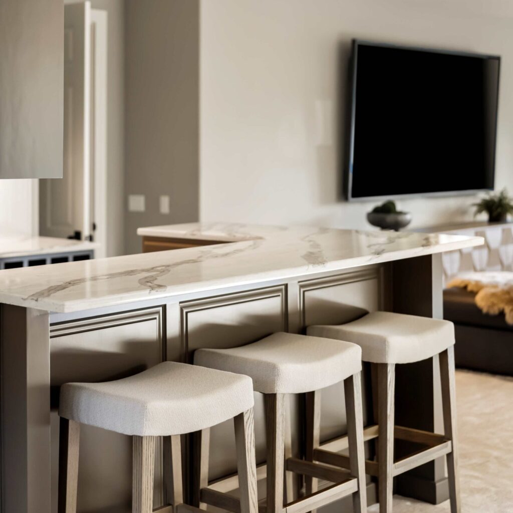 modern kitchen bar area featuring a curved marble countertop with four beige cushioned stools underneath. Neutral tones dominate the space, with a wall-mounted flat-screen TV and minimalistic decor visible in the background. Illustrating basement remodel. 
