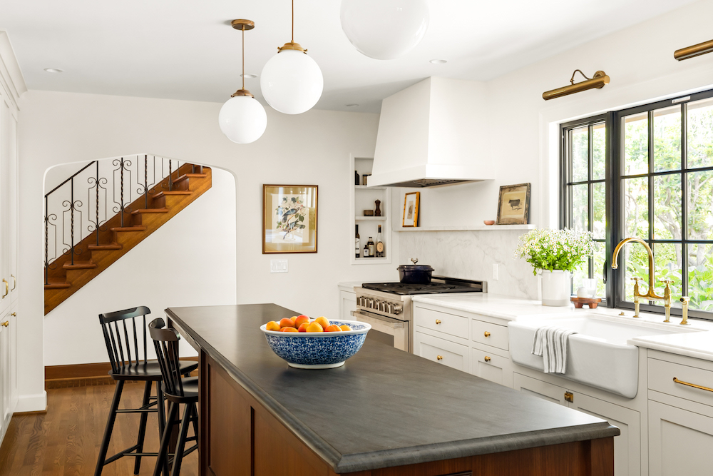 Bright kitchen: dark island, white cabinets, large window, pendant lights, and a staircase