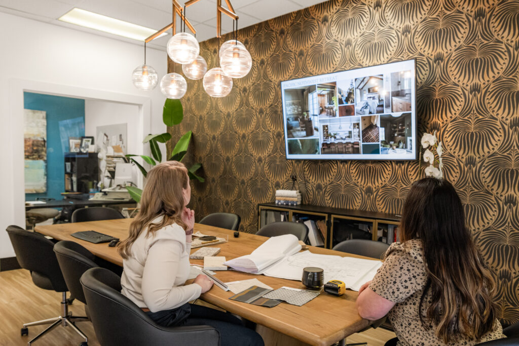  Two women seated at a conference table in a modern office, reviewing architectural plans and images displayed on a wall-mounted screen. The room features patterned wallpaper, hanging globe lights, and various office supplies, indicating a collaborative design or planning meeting.