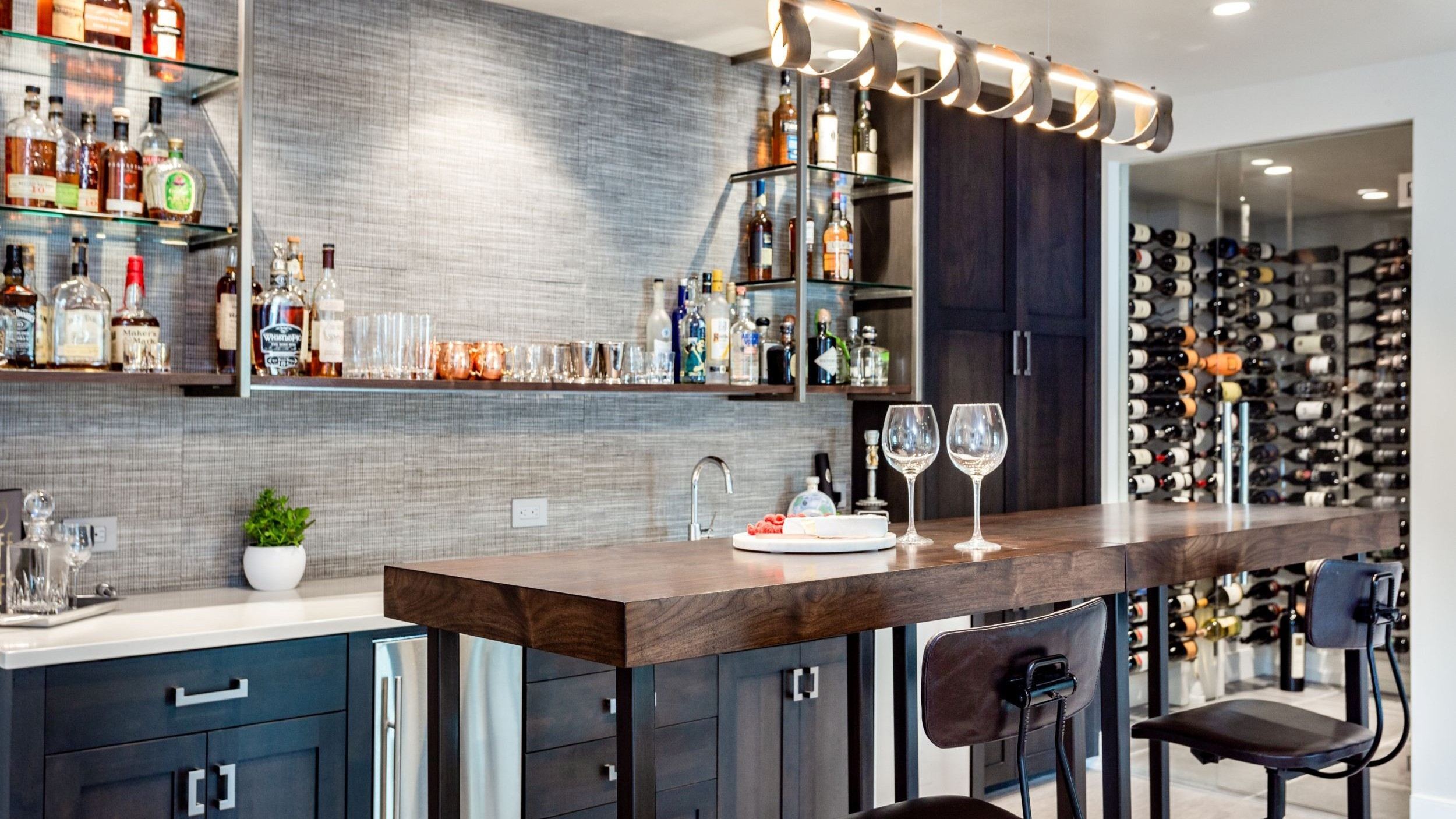 Modern home bar with dark cabinetry, wood countertop island, glass shelving stocked with liquor, under-cabinet sink, two bar stools, and a walk-in wine cellar with wall-mounted racks in the background. Illustrating basement remodel. 
