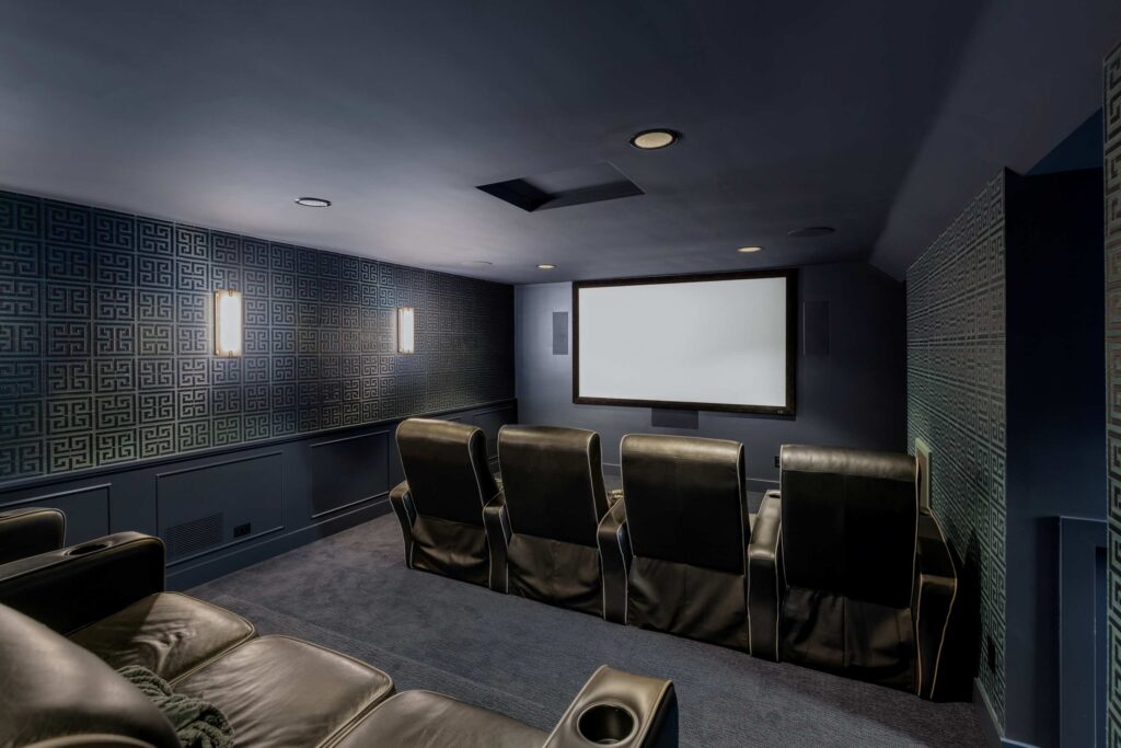 home theater room featuring a large blank projection screen and two rows of black leather reclining seats with cup holders. The room has dark blue walls with patterned wallpaper, recessed ceiling lights, and built-in speakers on either side of the screen, creating a cozy entertainment space. Illustrating basement remodel. 