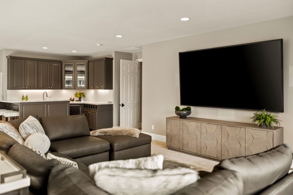 Modern living room and kitchen area showcasing a large flat-screen TV mounted on a light-colored wall above a textured wooden console with decorative plants. The space features a dark leather sectional sofa with patterned pillows, taupe cabinetry with under-cabinet lighting, and an open doorway leading to another room. Illustrating basement remodel. 