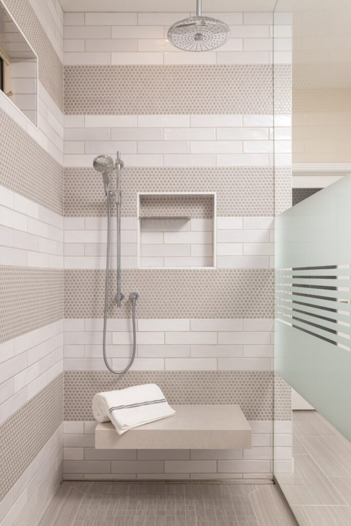 Remodeled shpwer with light/white tiles, bench with rolled towels, and glass enclosure to help illustrate how to survive a bathroom remodel.