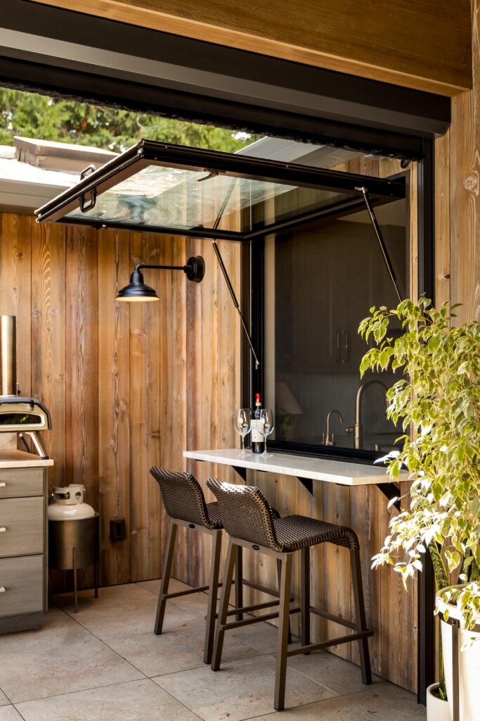 Outdoor living space with small bar seating by large window that opens to the indoor kitchen to help illustrate Does Outdoor Living Space Add Value?