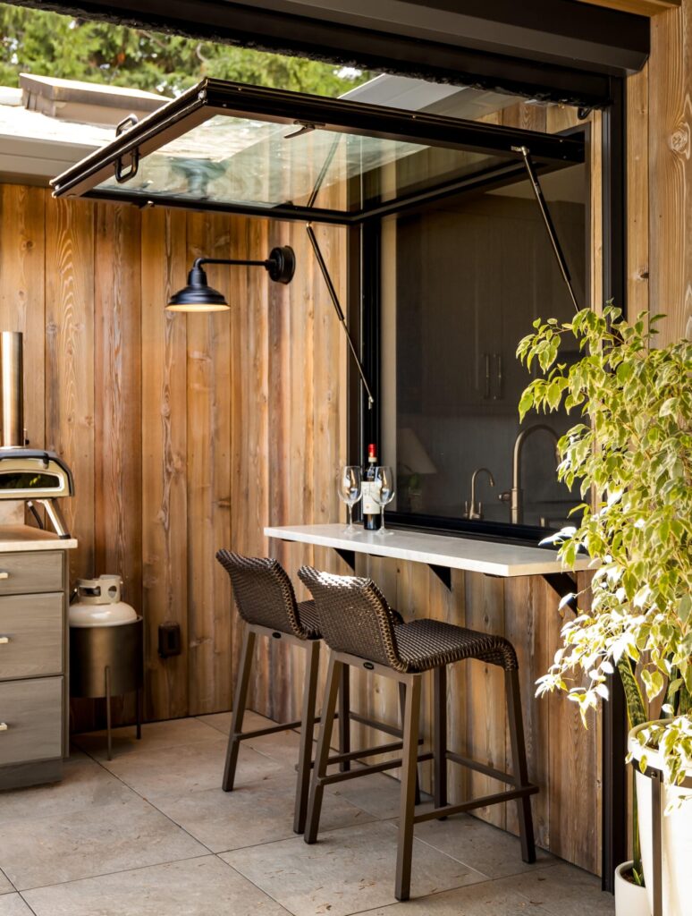 Outdoor living space with small bar seating by large window that opens to the indoor kitchen to help illustrate Does Outdoor Living Space Add Value?